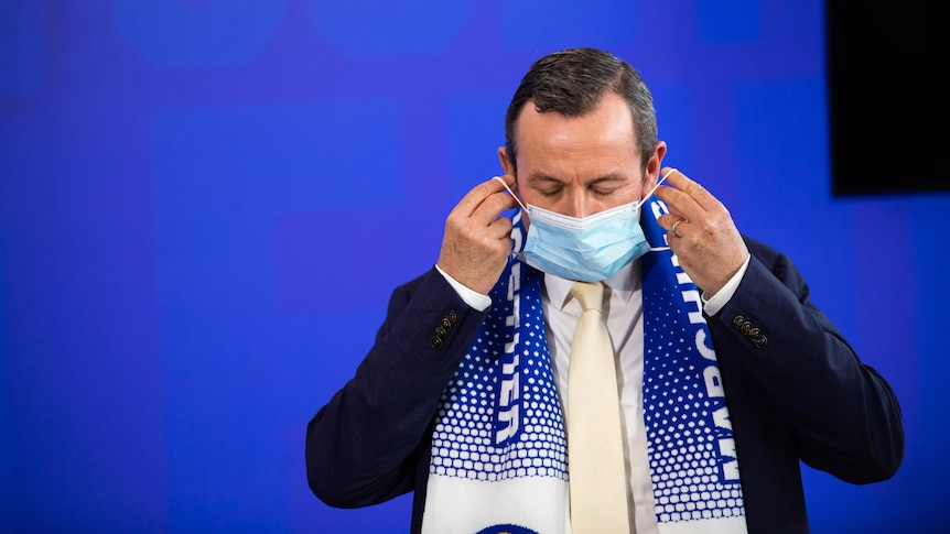 Premier Mark McGowan, wearing a suit, puts a mask on with his eyes closed.