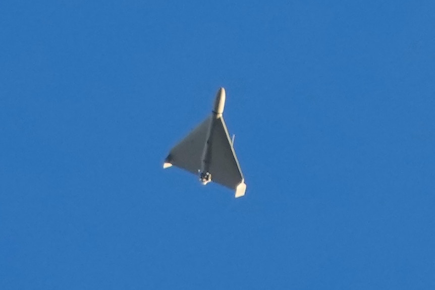 A white, triangular drone with a pointed warhead on its nose can be seen flying through the sky in a zoomed-in photo.