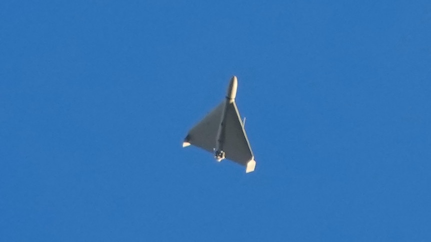 A white, triangular drone with a pointed warhead on its nose can be seen flying through the sky in a zoomed-in photo.