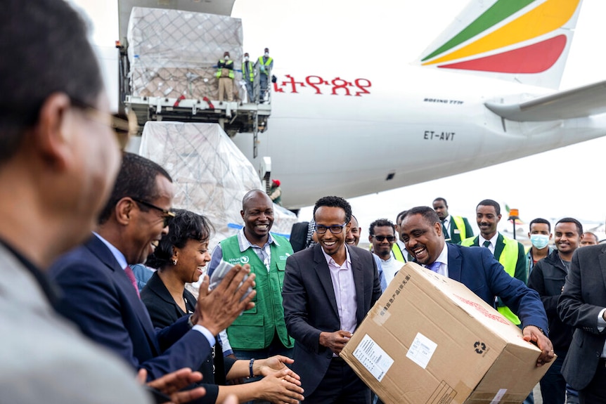 On the tarmac, you see a smiling crowd of Ethiopian officials holding a large box of medical supplies as a jet unloads behind.