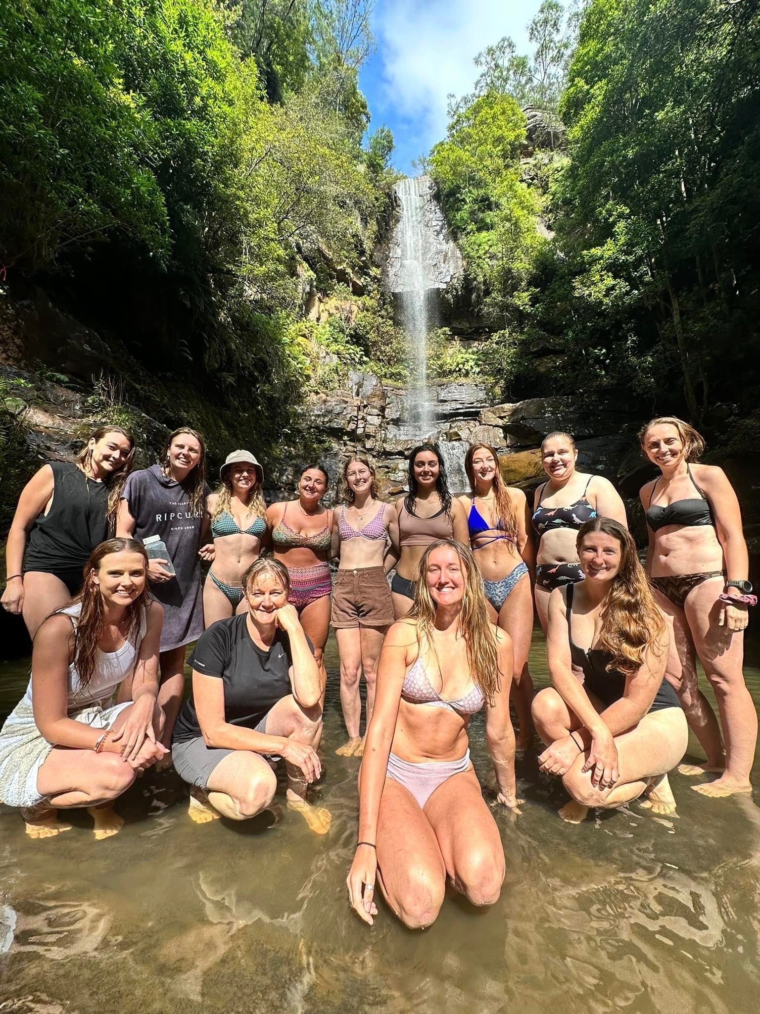 Women wearing swimmers stand at the base of waterfall 