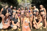 Women wearing swimmers stand at the base of waterfall 