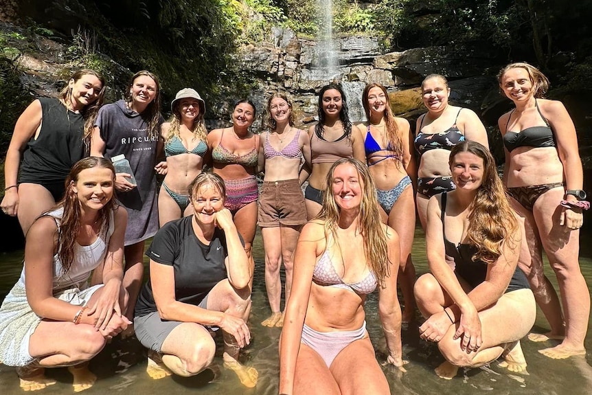 Women wearing swimmers stand at the base of waterfall 