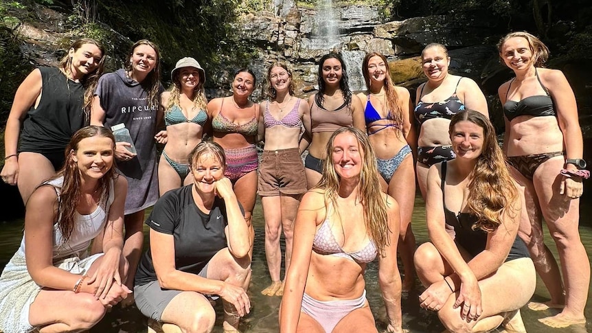 Women wearing swimmers stand at the base of waterfall 