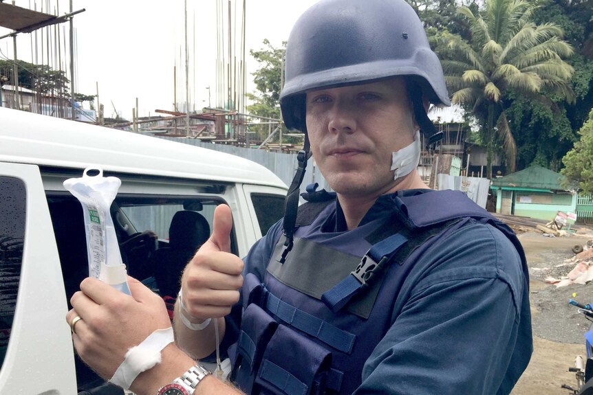 Harvey signals thumbs up, wearing a flak jacket and helmet with a dressed wound on his neck