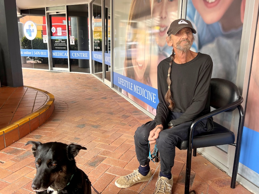 Man sitting outside a doctors surgery with a dog on the lead not smiling.
