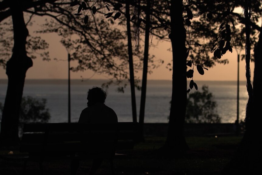 A person sits on a park bench at dusk overlooking the ocean they are silhouetted.