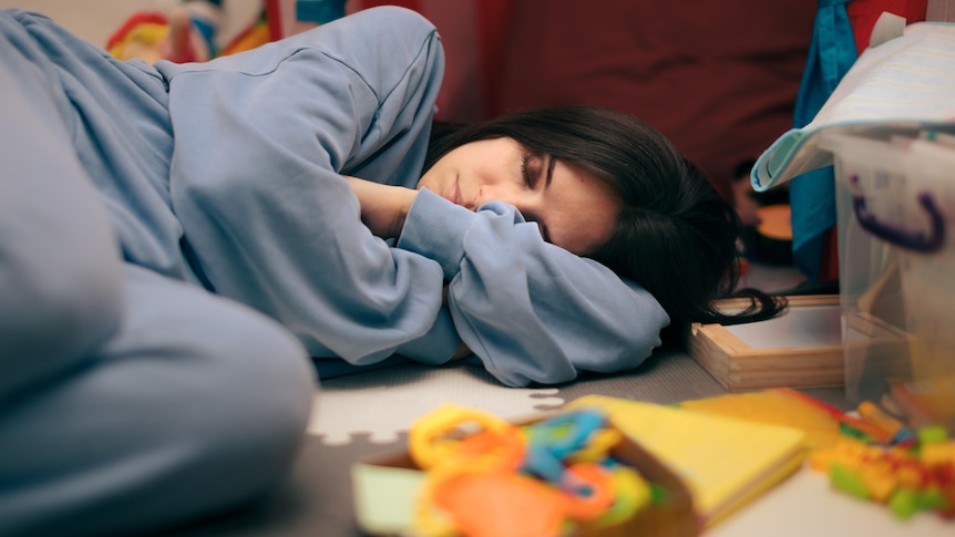 A woman in a blue tracksuit lies curled up on the floor with her eyes closed surrounded by toys and building blocks