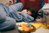 A woman in a blue tracksuit lies curled up on the floor with her eyes closed surrounded by toys and building blocks