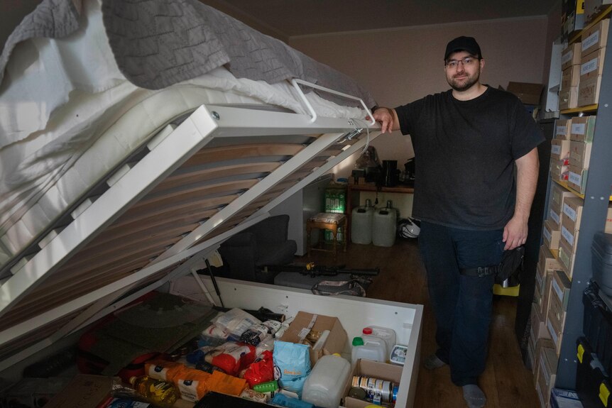 A man shows food items stored under his bed, lifting up the frame and mattress. 