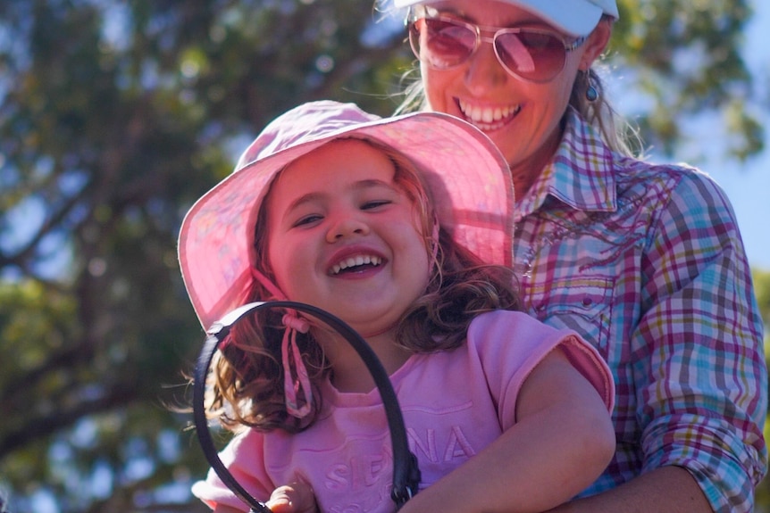A little girl with a pink hat, with a big grin, sitting on a horse in front of a female adult rider who is laughing.