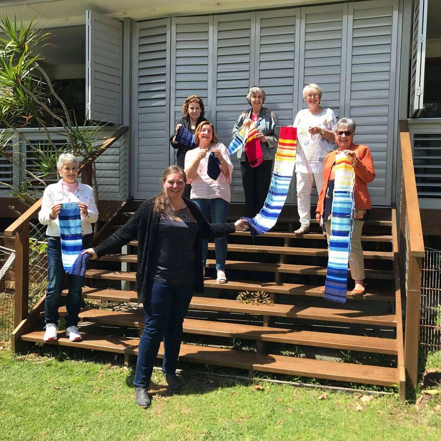 CEO Brooke Prentis stands on the steps of a house with six women, all smiling and holding scarves of coloured stripes