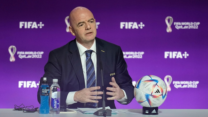 Gianni Infantino sits at a table with a ball in front of him with his hands stretched out