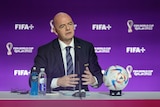Gianni Infantino sits at a table with a ball in front of him with his hands stretched out