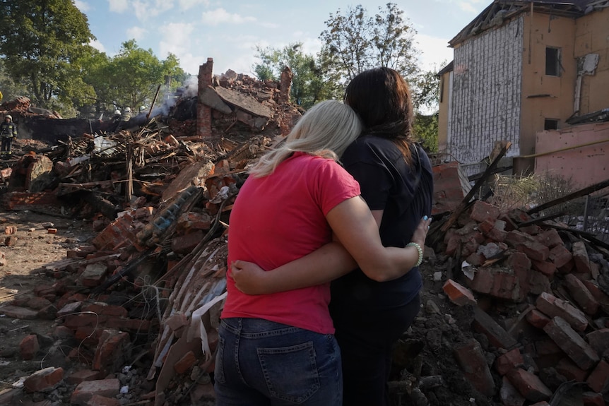 Ukrainian women hug in front of a destroyed building