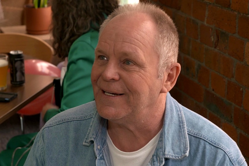 A man in a blue jean shirt sits at a table in a restaurant and smiles.