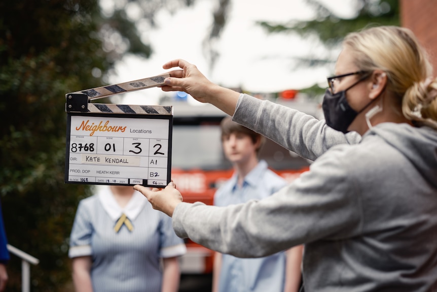 A crew member wearing a mask holds a clapperboard on the set of Neighbours.
