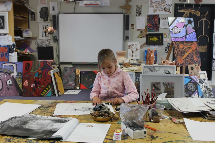 A girl in a pink jumper draws on a piece of paper with charcoal surrounded by colourful canvases and art