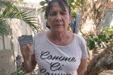 A mid shot of an Indigenous woman in a grey shirt standing in a backyard garden holding a cashless welfare card.
