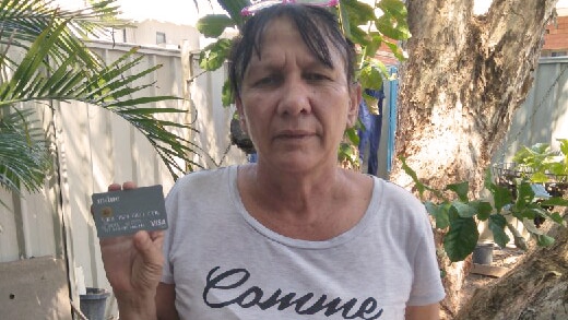 A mid shot of an Indigenous woman in a grey shirt standing in a backyard garden holding a cashless welfare card.