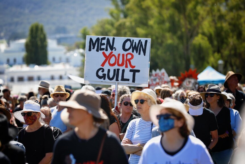 A woman in a crowd holds a white sign reading MEN, OWN YOUR GUILT