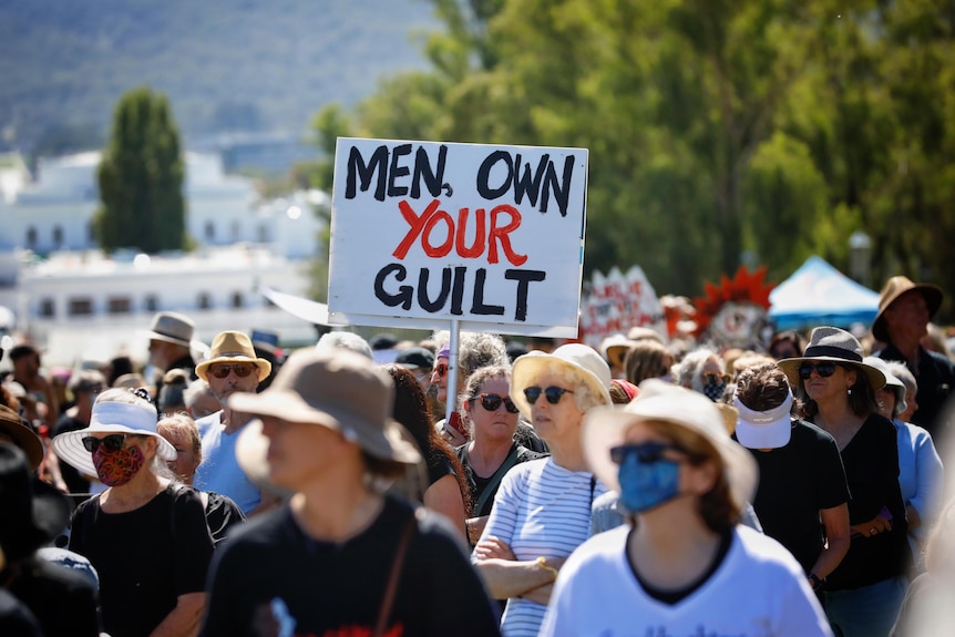 A woman in the crowd holds a white sign that says MEN, OWN YOUR GUILT.