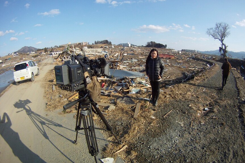 Woman standing amid debris in front of a camera on a tripod.