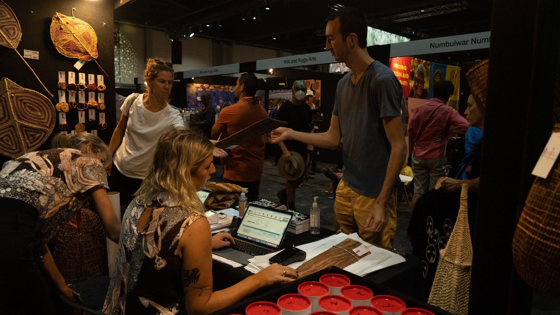 Two people stand at a desk to pay for an artwork, as an art fair goes on in the background. 