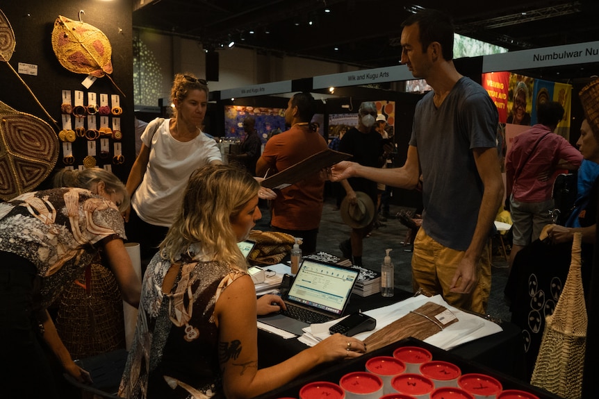 Two people stand at a desk to pay for an artwork, as an art fair goes on in the background. 