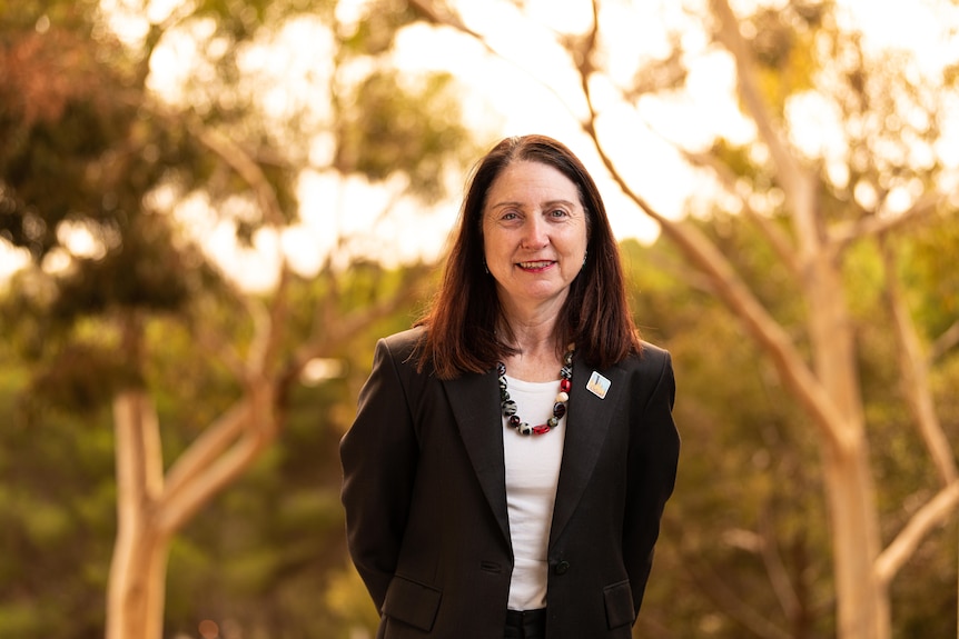 Woman with black blazer and beads around her neck smiling at the camera with trees in the background