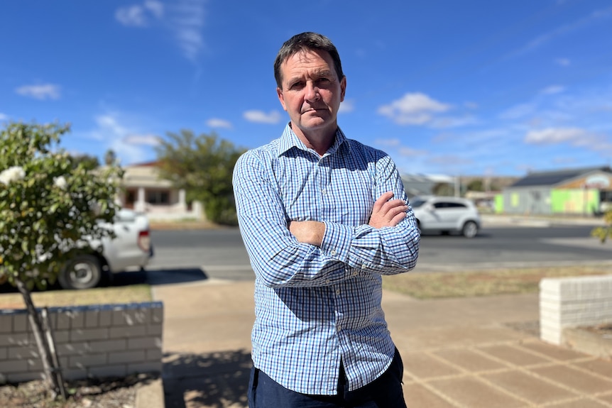 A frowning man wearing a shirt with his arms crossed in front of a street. 