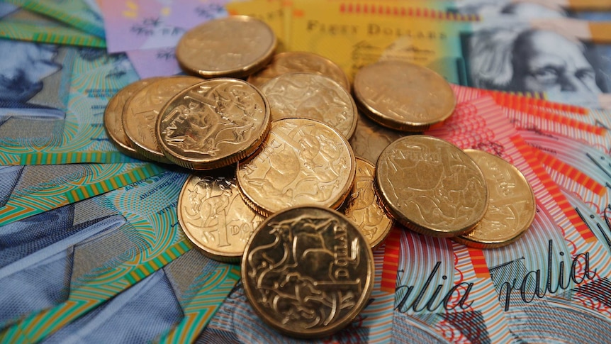 A pile of Australian one dollar coins sits on banknotes of various denominations.