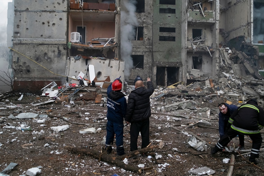 Two people stand looking up at a destroyed apartment building. Workers to the right shift rubble.