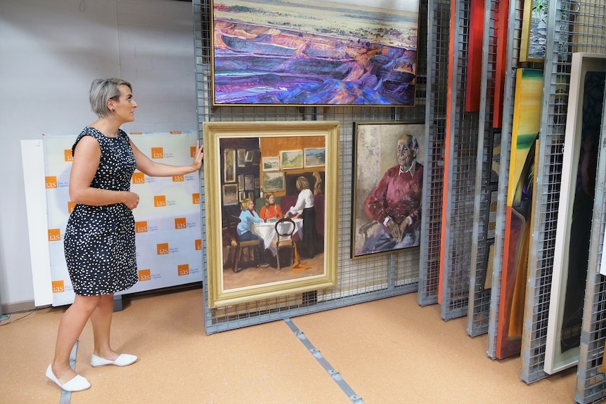 A woman pushing a sliding rack of artwork in a store room, colourful 2D artworks on display.