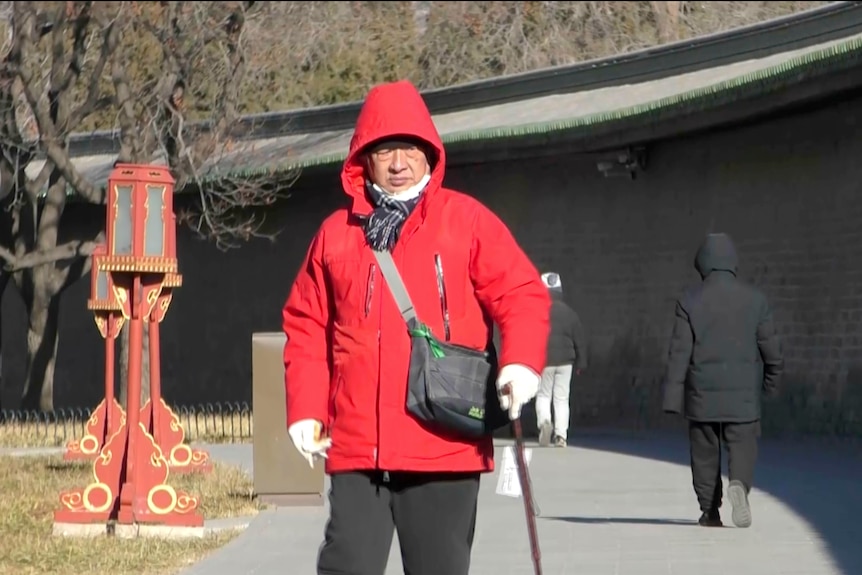 Пожилой мужчина с тростью на улице в красной куртке и кожаной сумке через плечо.