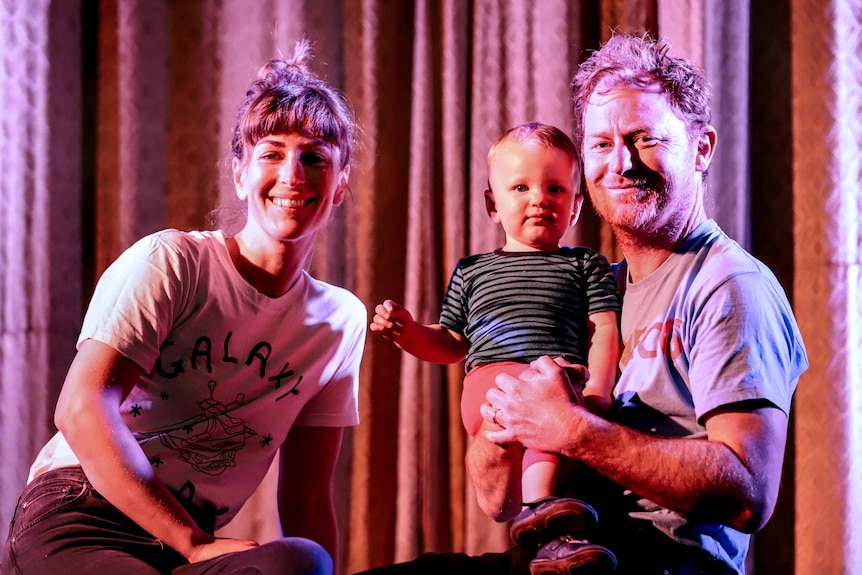 A young woman in a white t-shirt sits on a stage next to a man in a blue t shirt holding a baby with a large pink curtain behind