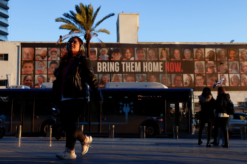 People at a bus stop walk past a building with a large poster with pictures of dozens of people, it reads 'bring them home now'