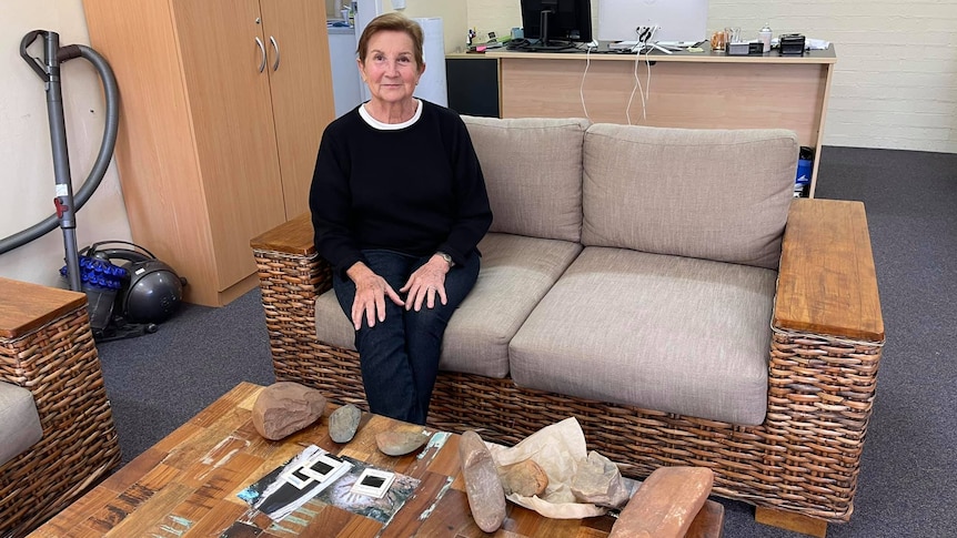 An older lady sits on a lounge with Aboriginal artefacts on the coffee table in front of her.