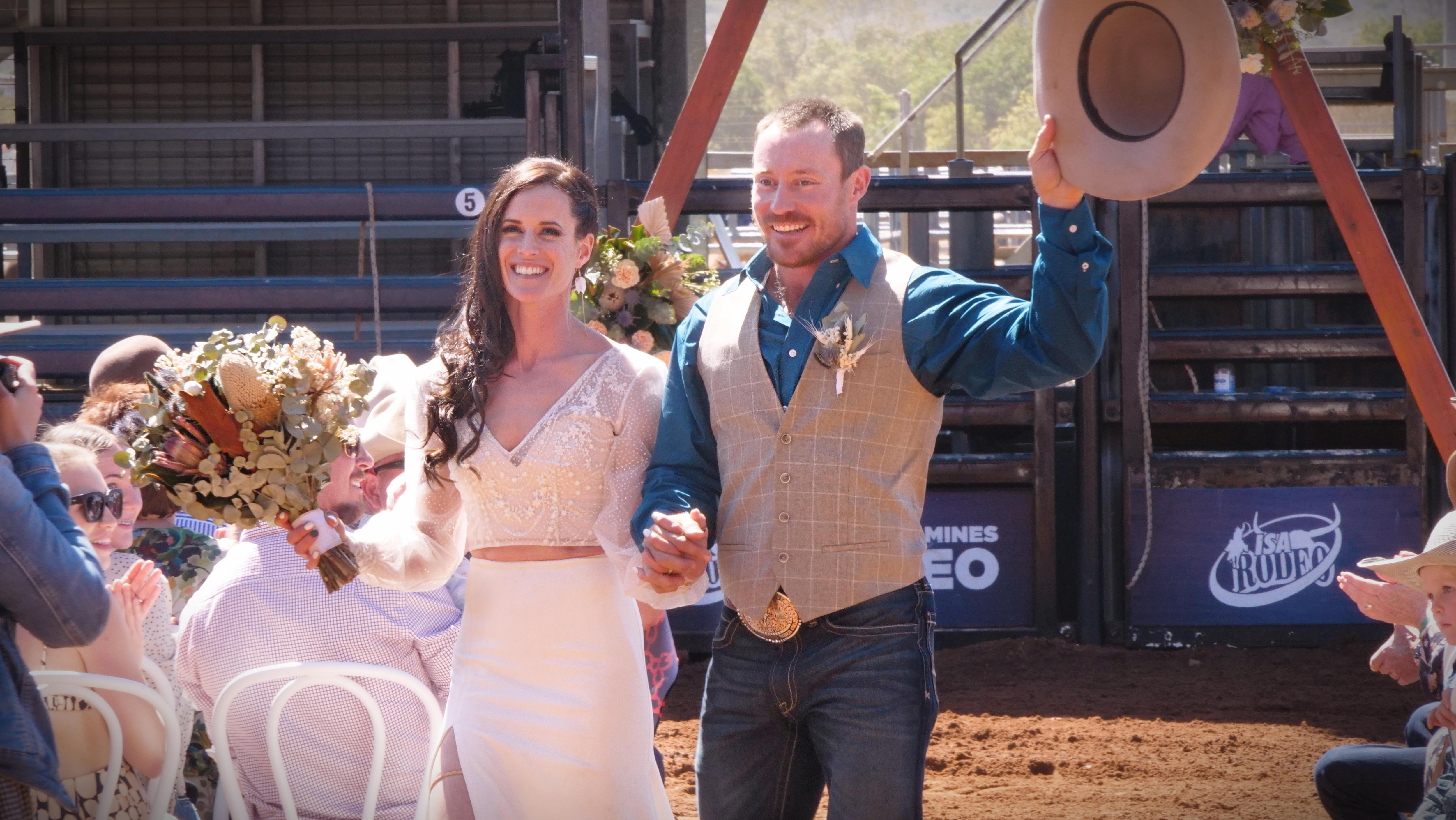 A bride in a white dress with long brown hair walks alongside her groom wearing a blue shirt and cream vest holding a cowboy hat
