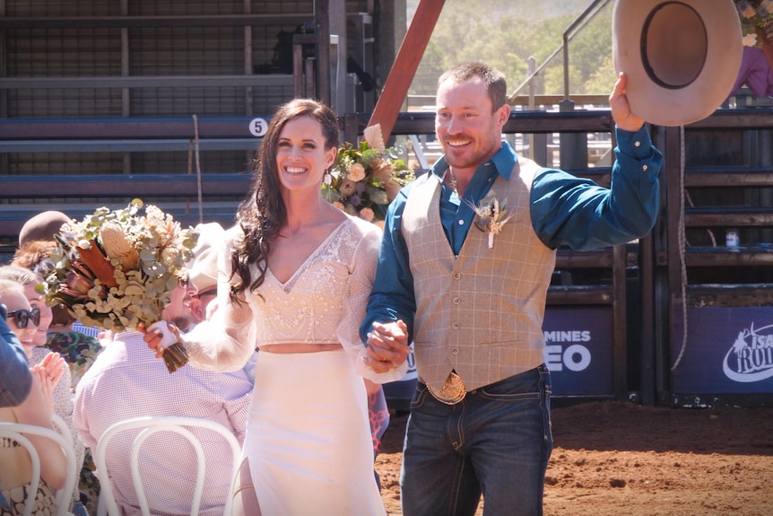 A bride in a white dress with long brown hair walks alongside her groom wearing a blue shirt and cream vest holding a cowboy hat