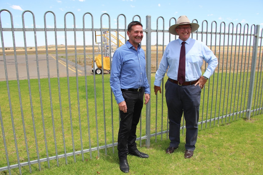 Two white men smiling at a camera, standing in front of the fence at the Broken Hill aiport.