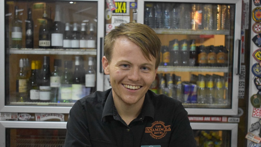 A young man with blonde hair smiles at the camera.  There are bottles of alcohol behind him.