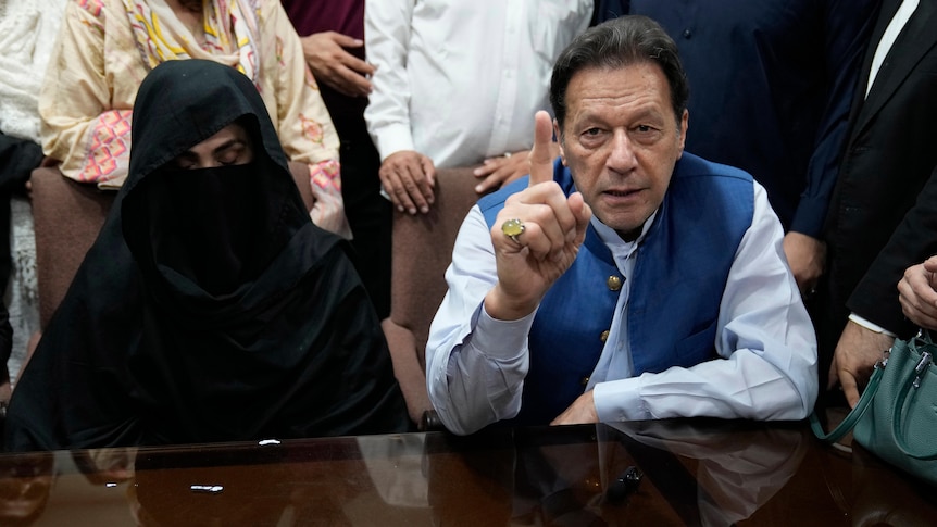 A man in a blue shirt and vest sits next to a woman in dark black conservative Islamic garb behind a table.