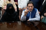 A man in a blue shirt and vest sits next to a woman in dark black conservative Islamic garb behind a table.