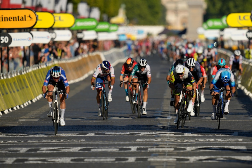 A cyclist on the far left of the picture wins a sprint to win a Tour de France stage, just ahead of two riders on the far right.