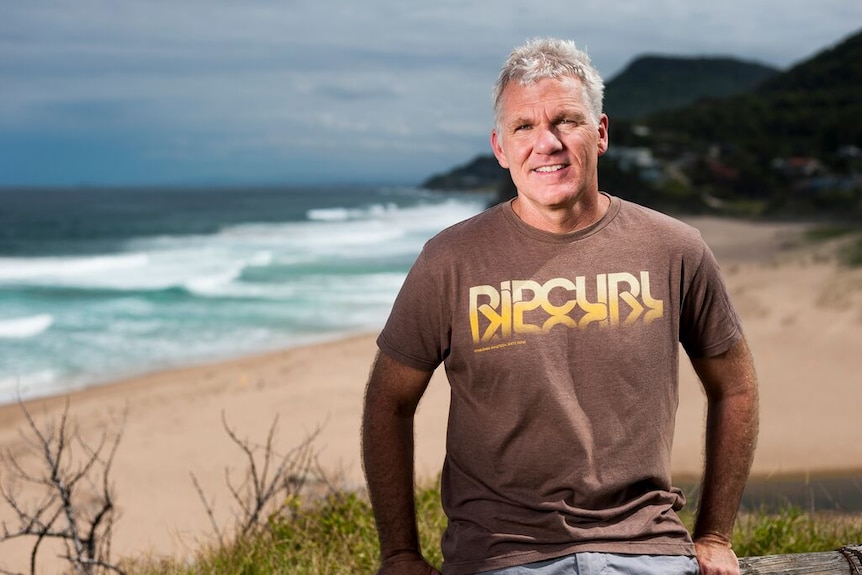 Middle-aged man in a brown t-shirt stands in front of a beach.