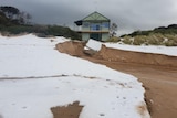 A damaged building on a beach with white hail on the sand in the foreground.