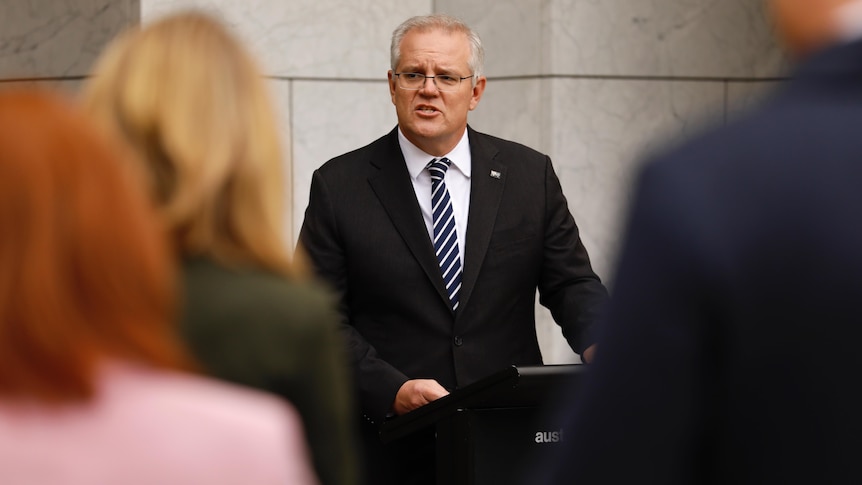 Morrison concentrates as he talks at a podium in front of a crowd of reporters