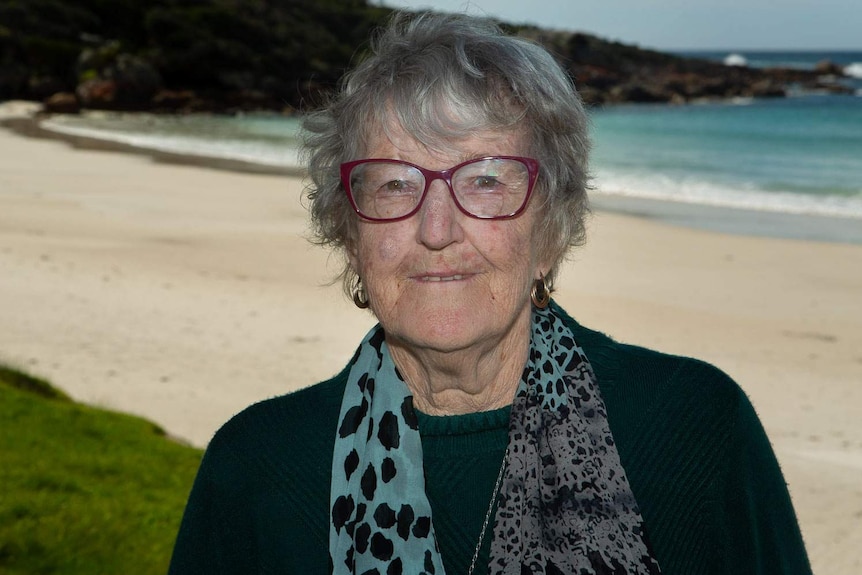 A woman with glasses stands on the foreshore of a harbour with sea lapping on beach behind.