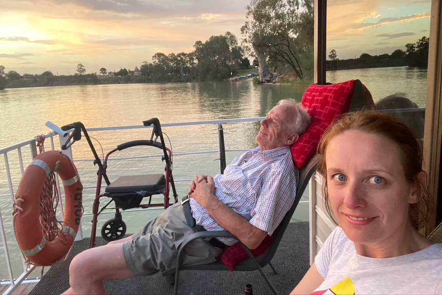 Elderly man sitting on a deck chair, smiling. A woman taking a selfie.
